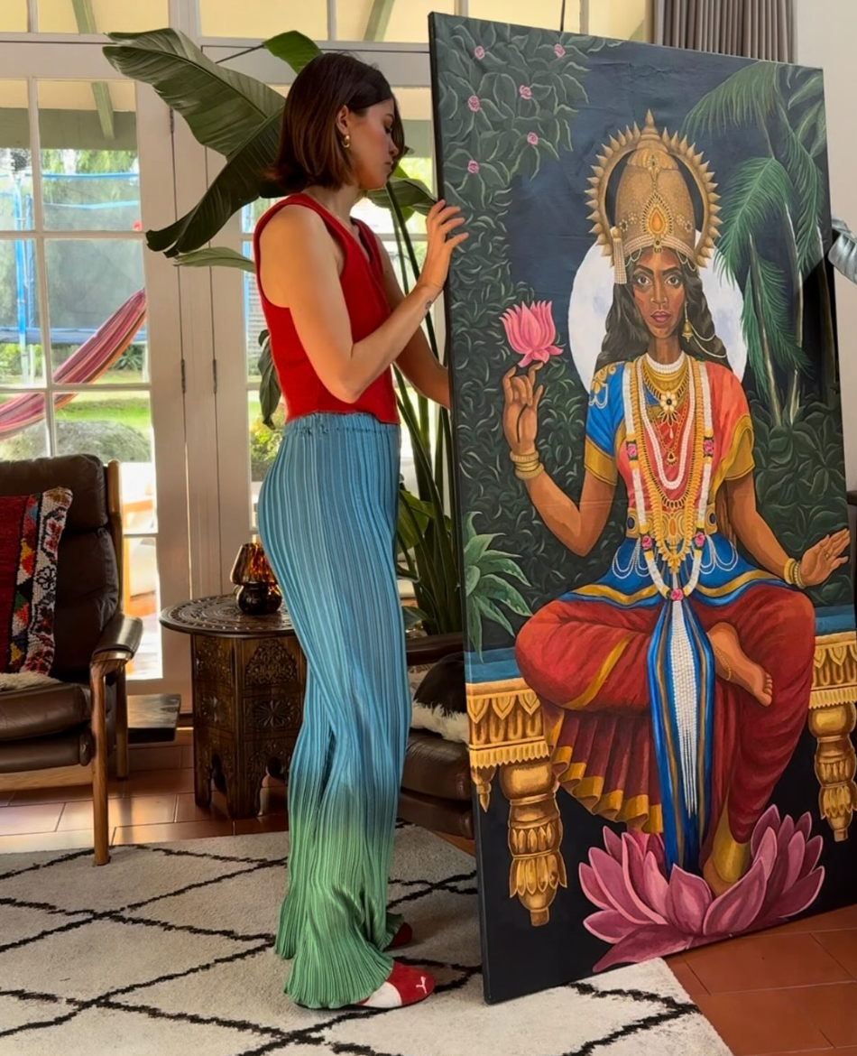 Woman standing next to a large painting of a deity in a room with plants and furniture.